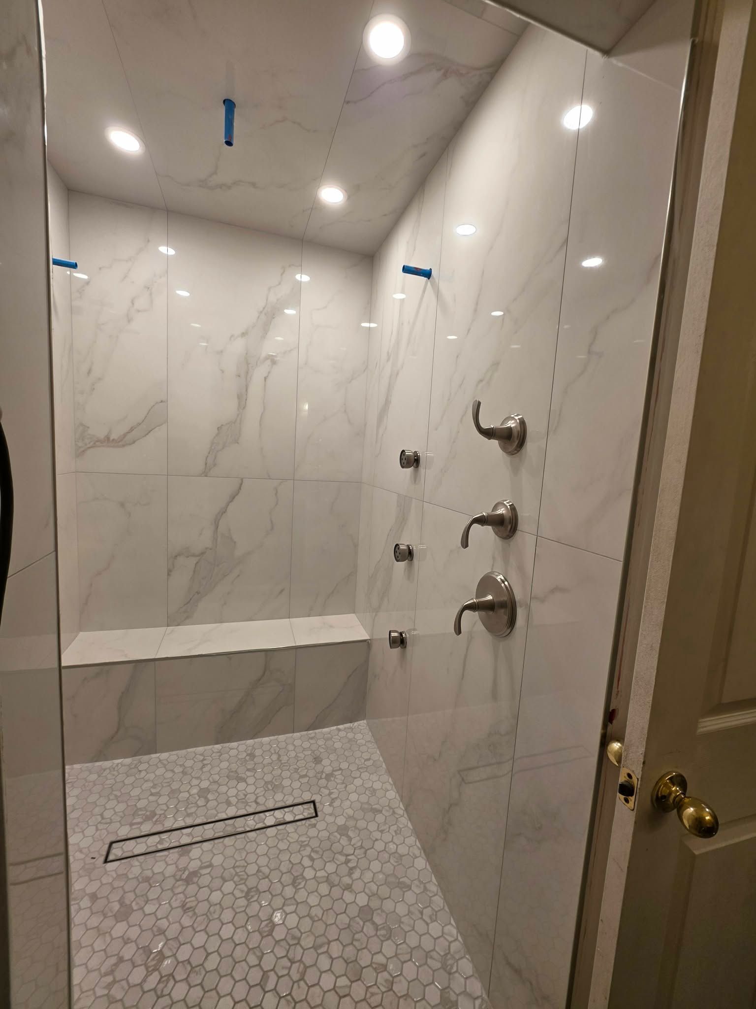 Small white tiled shower with marble-look walls, built-in bench, and chrome shower fixtures.