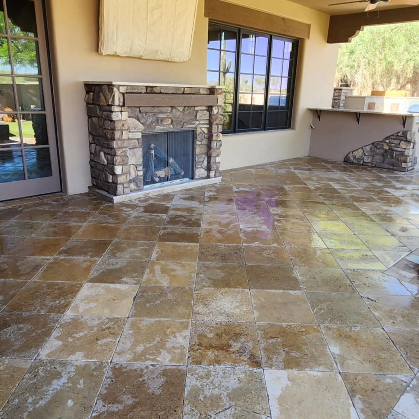 Covered stone patio with a fireplace, large windows, and wet tile floor.