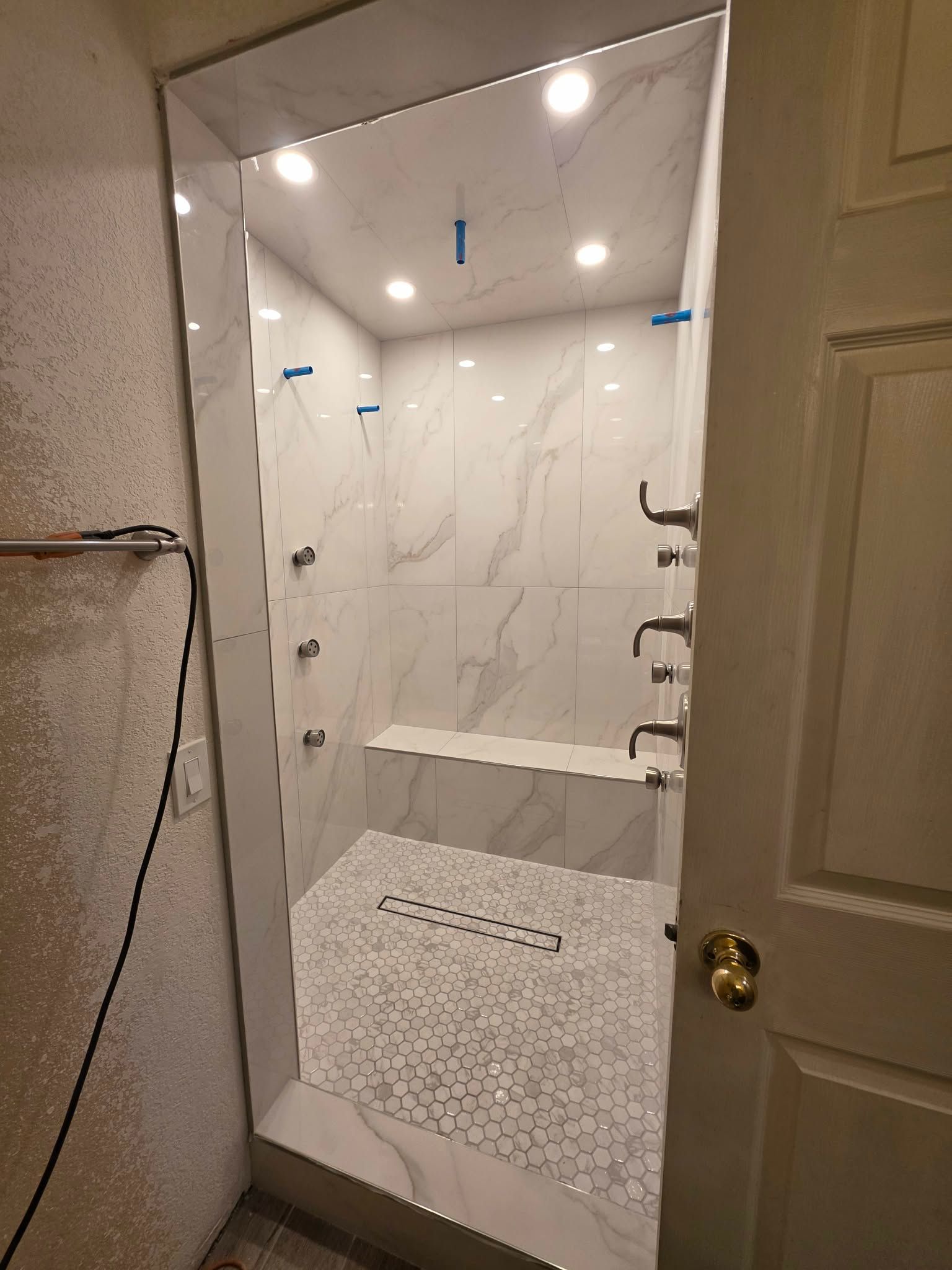 Bathroom doorway view of a marble-tiled shower with a bench, pebble floor, and partially open door