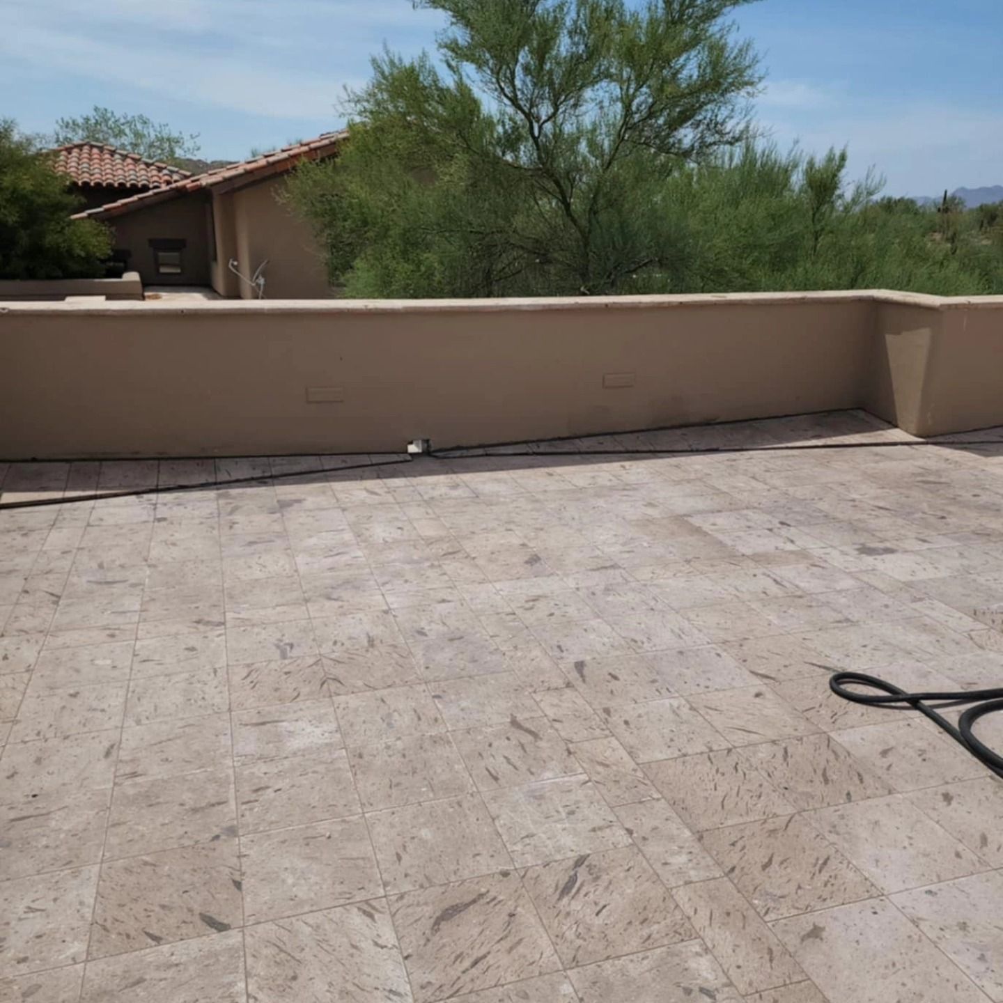 Empty stone patio with a beige wall, desert trees, and part of a garden hose in the corner.