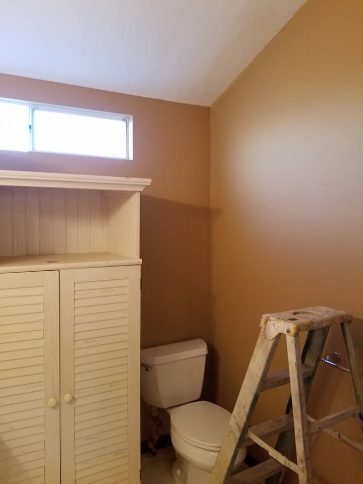 Small bathroom with toilet, white louvered cabinet, high window, and a step ladder under tan walls.