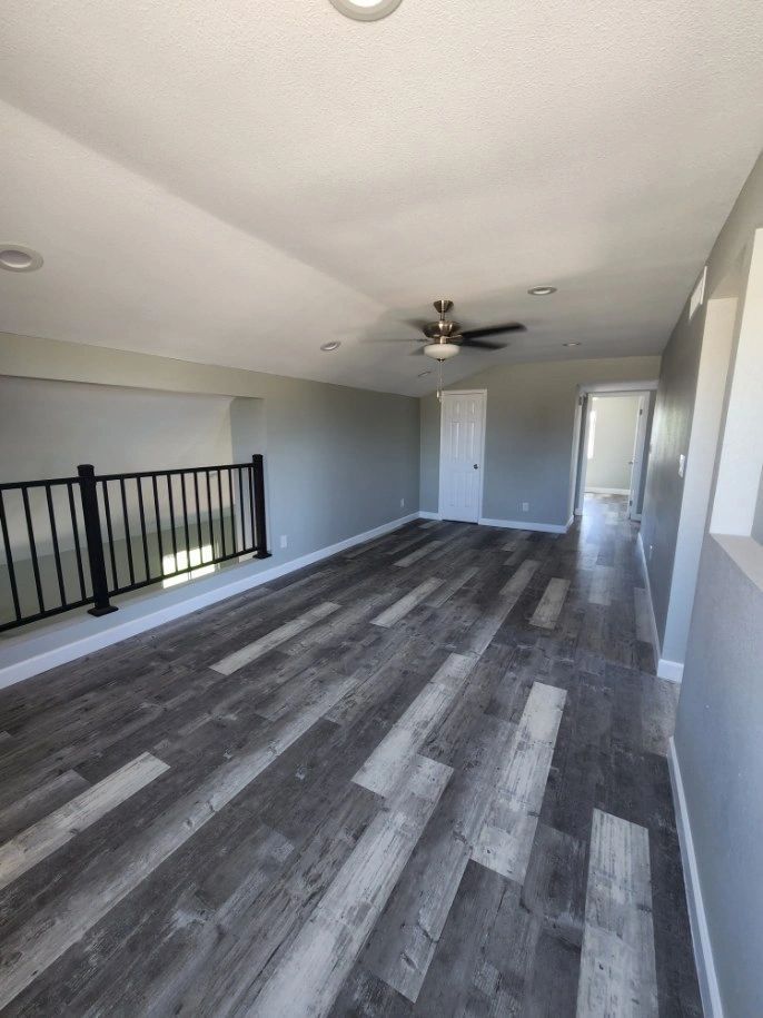 Empty upstairs hallway with gray wood floors, white walls, ceiling fan, and railing overlooking a lower level