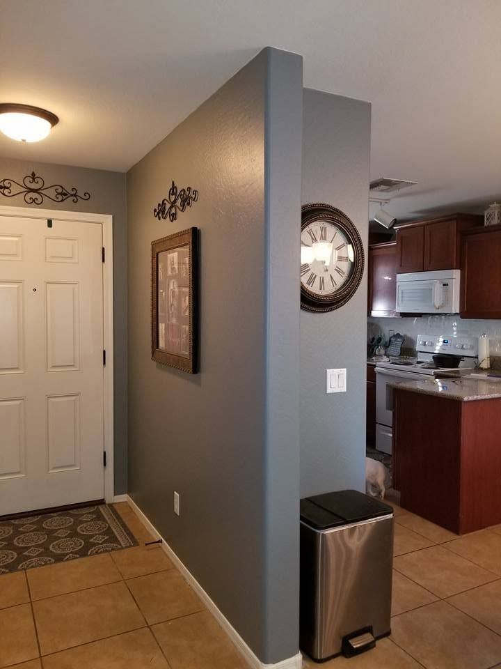 Apartment entryway with gray walls, white door, wall clock, and stainless trash bin near the kitchen.