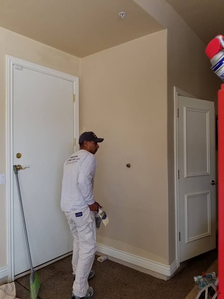 Person in white work clothes painting a beige room near a white door and baseboards.