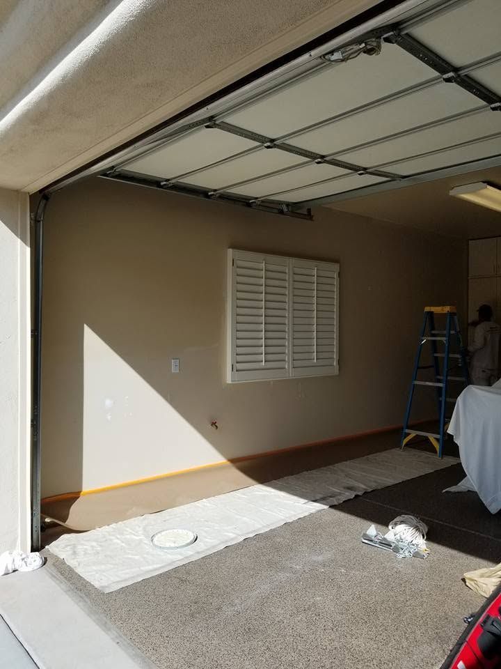 Open garage with raised door, beige wall, window shutters, and items inside on the floor.