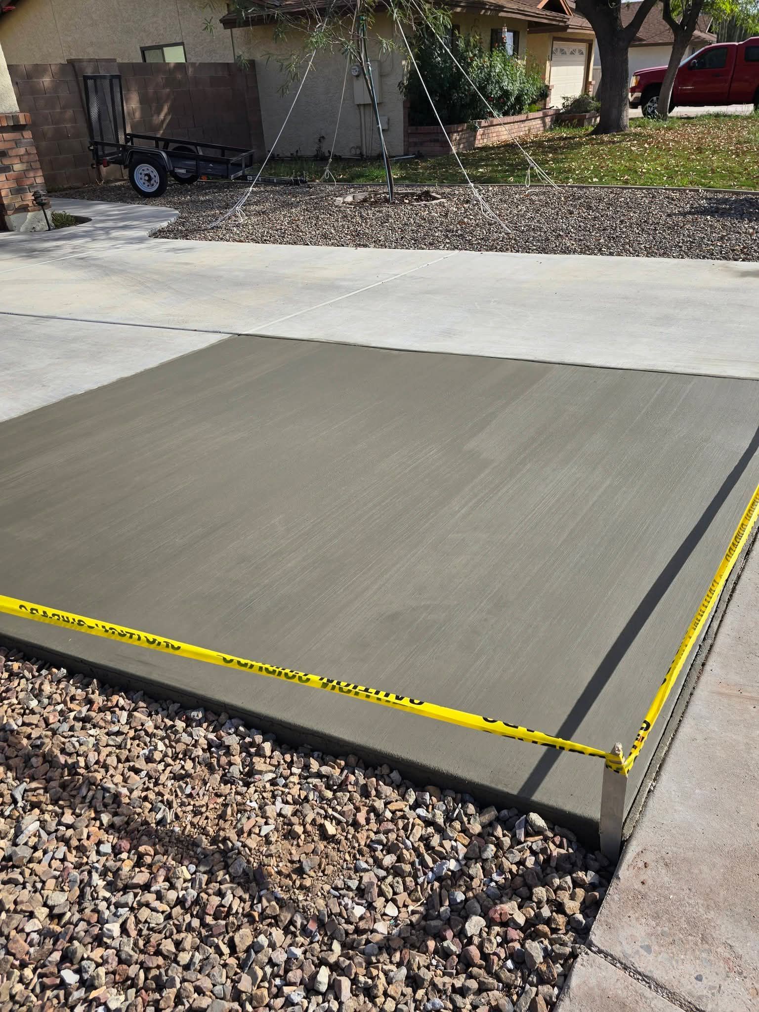 Fresh concrete driveway pad beside gravel and sidewalk, with yellow edge tape marking the perimeter