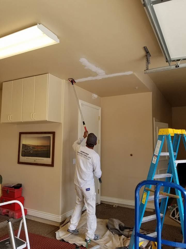 Painter rolling white paint on a garage ceiling beside a blue ladder and tan cabinets