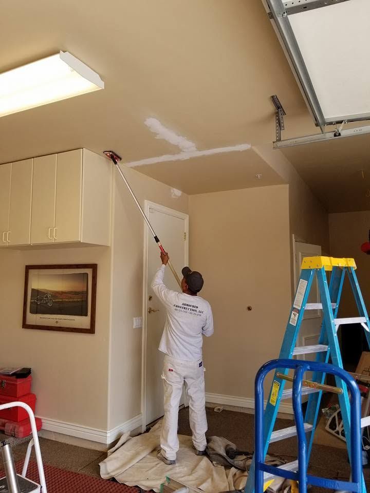 Painter in white painting a ceiling wall near a ladder inside a garage.