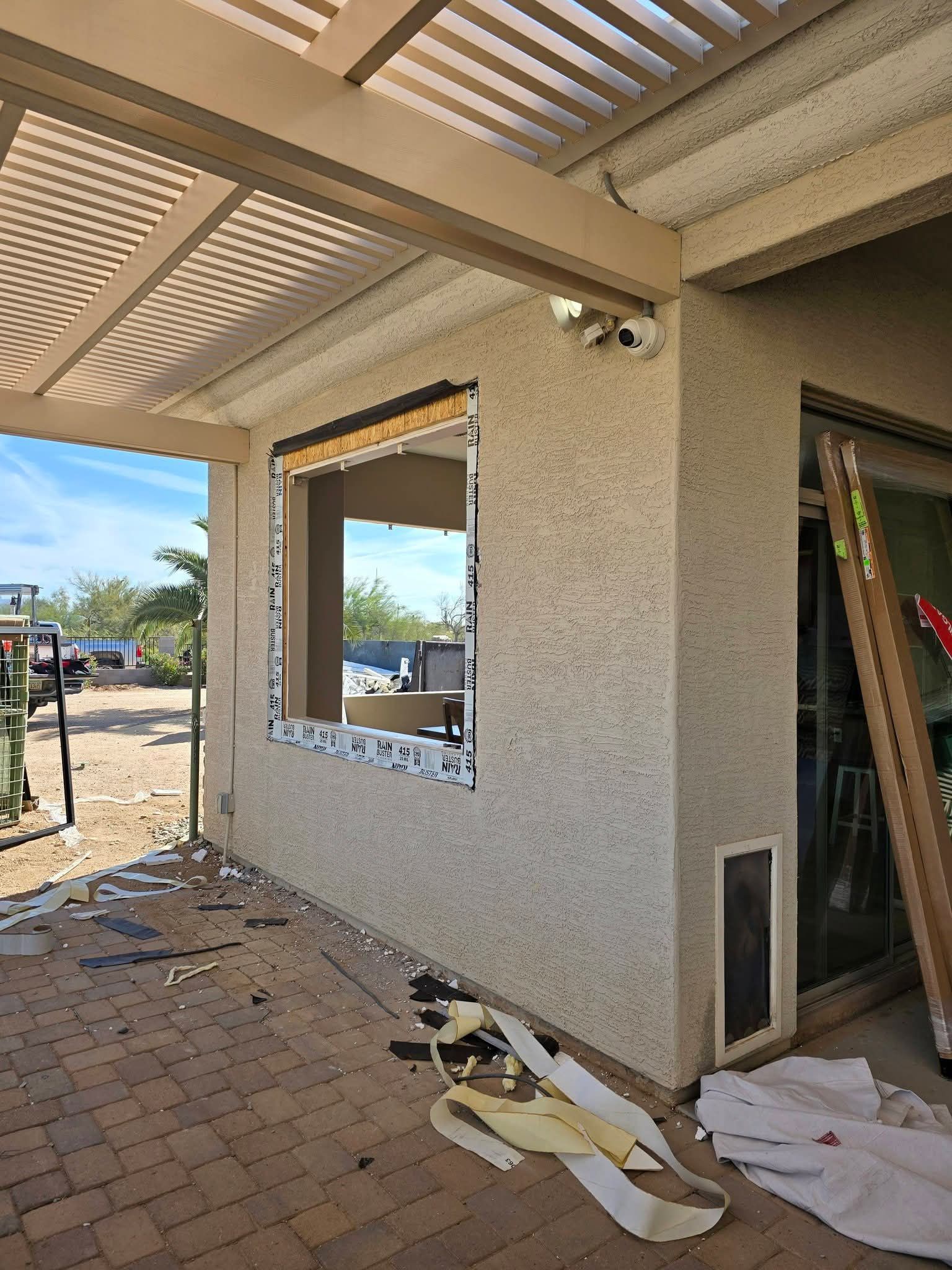 Outdoor patio under construction beside a stucco house, with tools and debris on the brick floor.