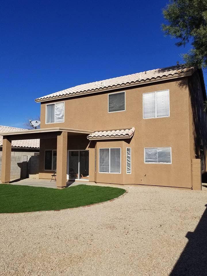 Two-story tan house with a covered patio, gravel yard, and blue sky.