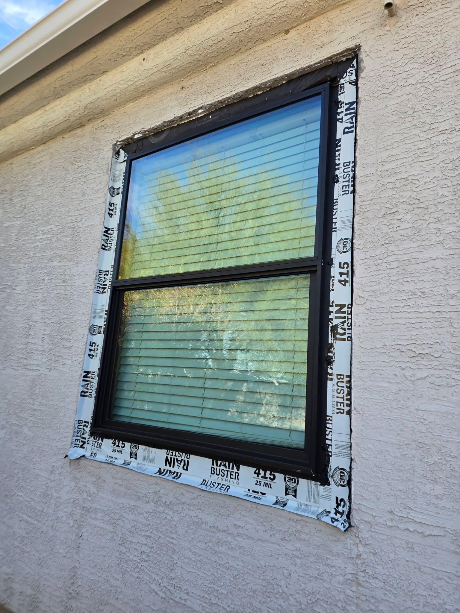 Single window with black trim in beige stucco wall, framed by exposed white house wrap