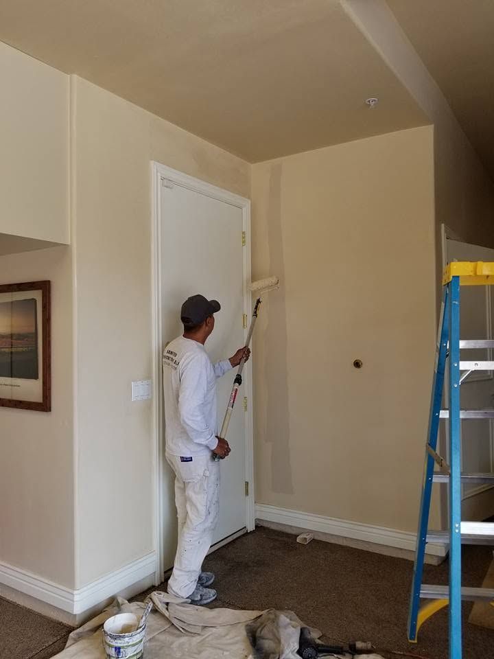 Painter painting a beige wall beside a white door in a room with a ladder and drop cloths.
