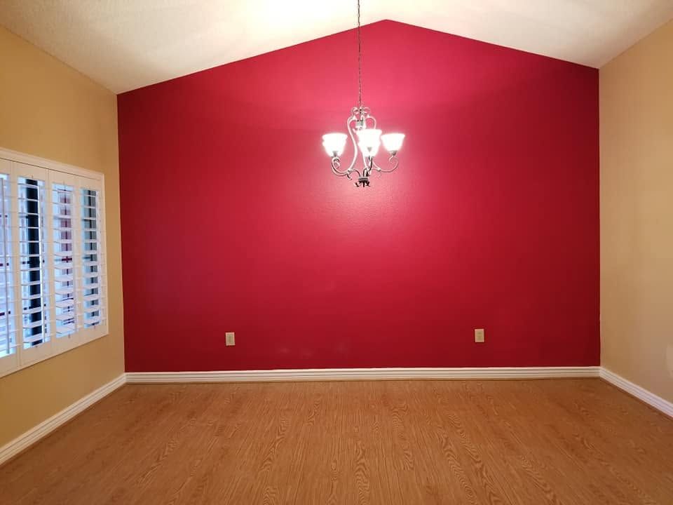 Empty room with red accent wall, beige walls, wood floor, windows, and a hanging chandelier.