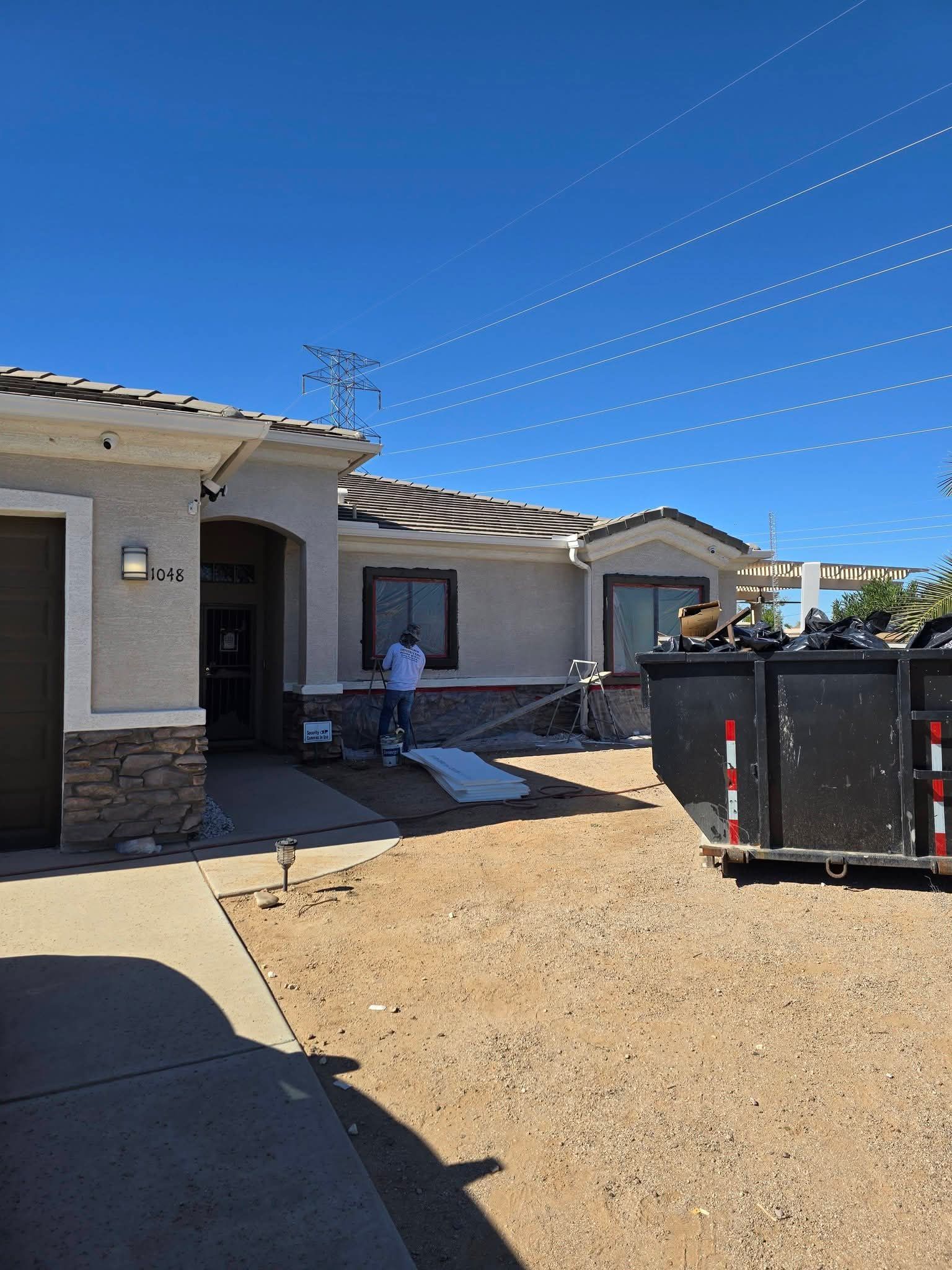 Single-story house exterior with a gravel yard and a large dumpster in front under a blue sky