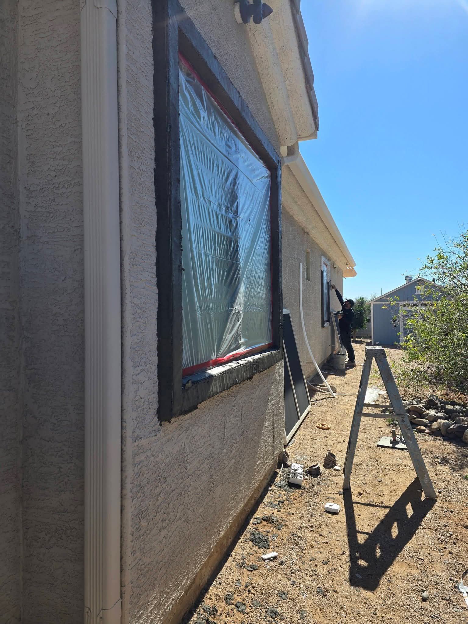 House exterior with stucco wall, reflective window, and a sunlit dirt path along the side yard.
