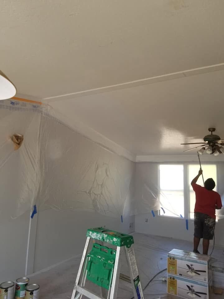 Person painting a white room ceiling, with a ladder, plastic-covered floor, and a window at the far end