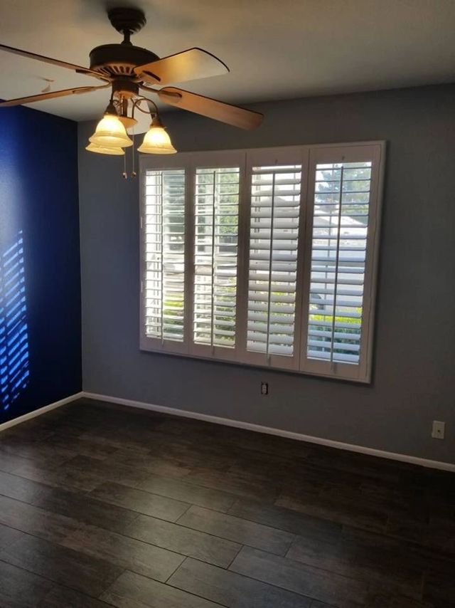 Empty room with dark wood floors, gray walls, white plantation shutters, and a ceiling fan light.