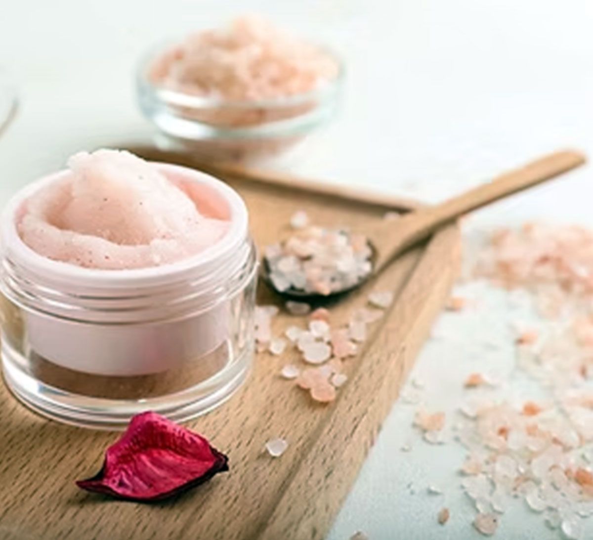 Pink body scrub in jar with wooden spoon, scattered salt crystals, and a rose petal on a wooden surface