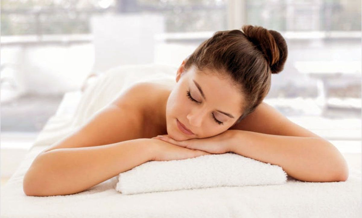 Woman resting her head on folded arms on a white massage table in a bright spa room