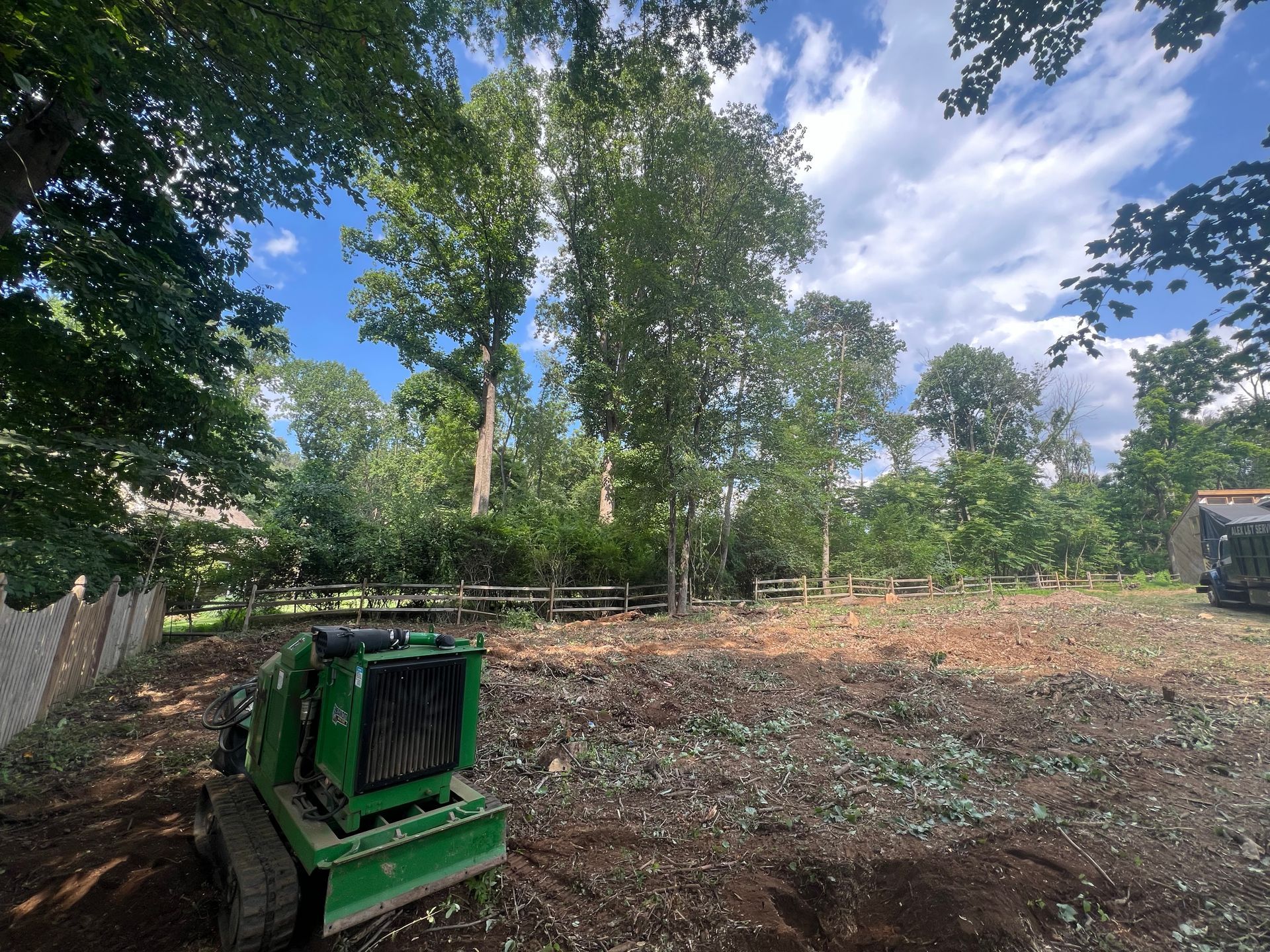 A green skid steer mower sits in a cleared, wood-chipped yard against a backdrop of trees under a blue, cloudy sky.