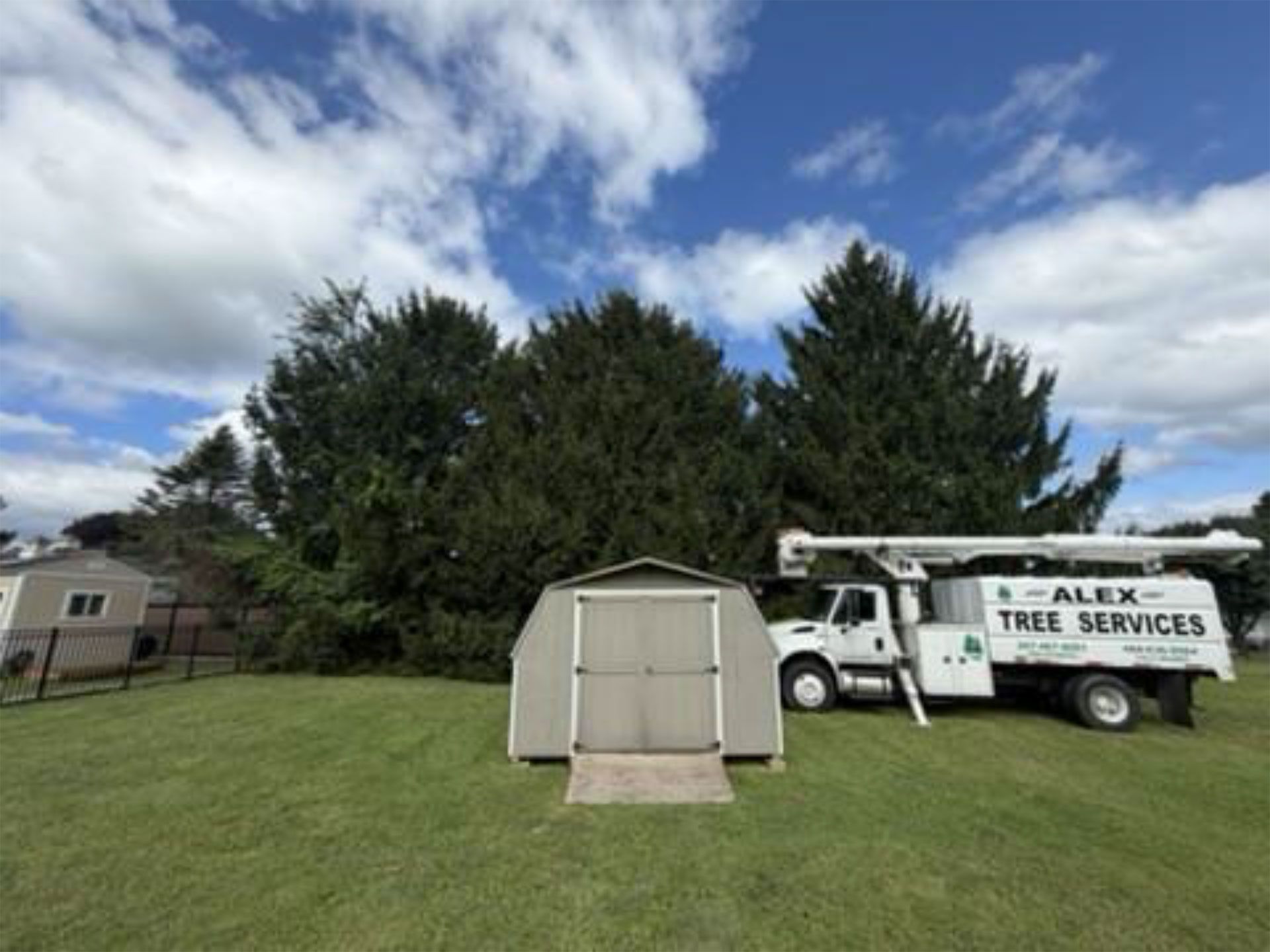 Tree service truck and trailer beside a small shed on a grassy lawn under a cloudy sky.