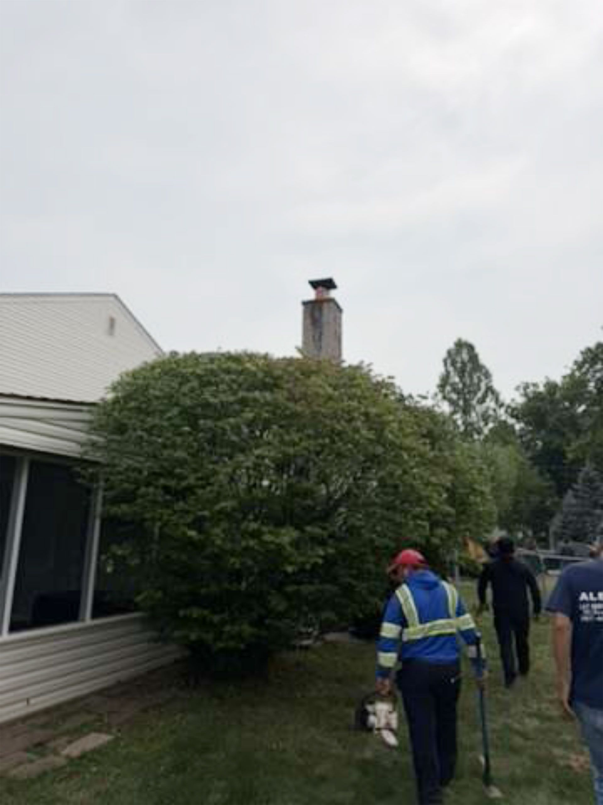 People in safety gear inspect a house with smoke coming from the chimney in a wooded area.