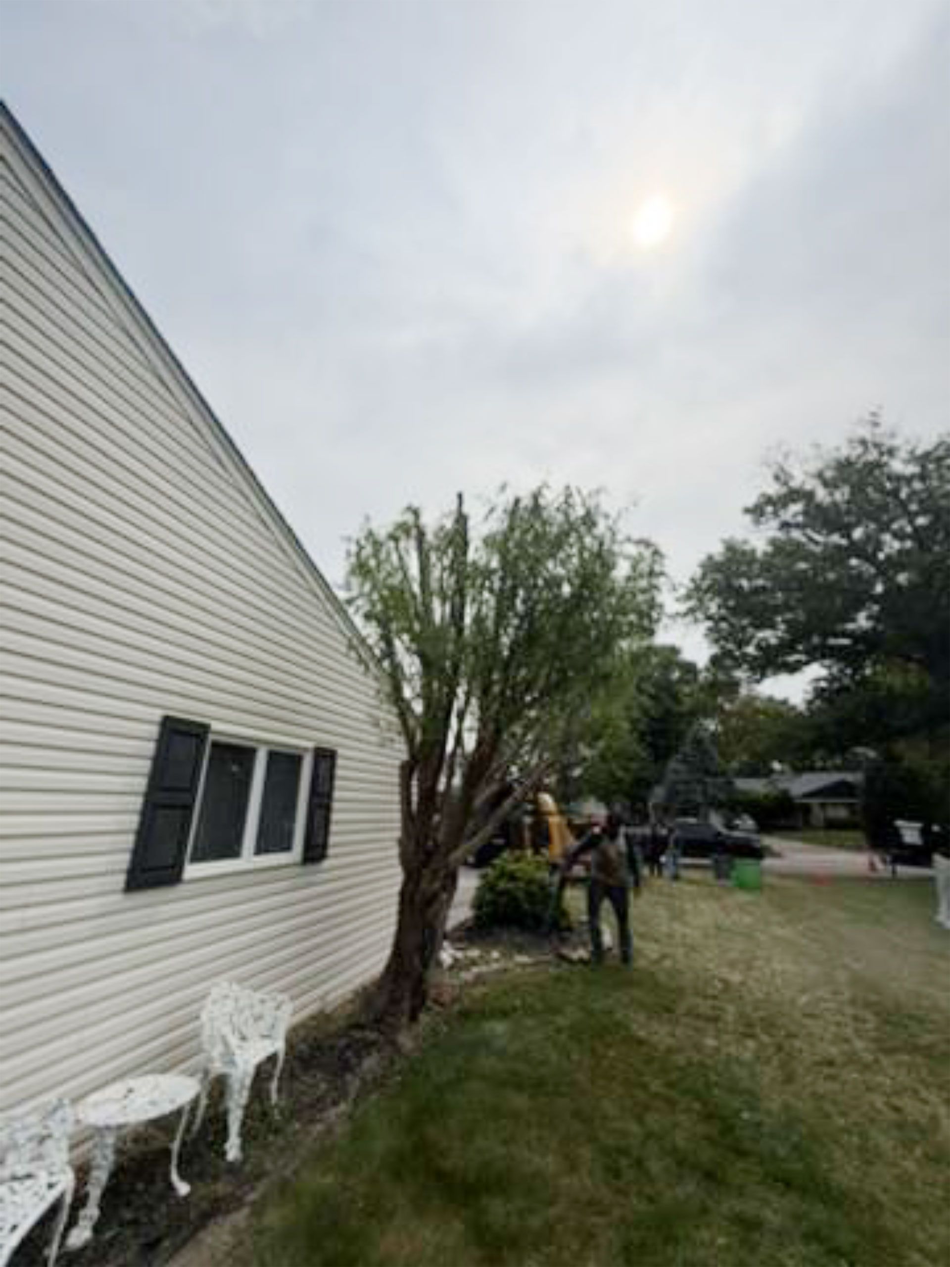 Person mowing grass beside a white house with a tree and outdoor chairs in the yard.