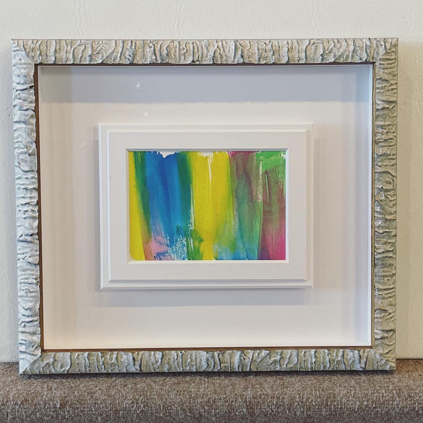 A small abstract painting in a white frame, centered in a larger textured frame; bright colors.