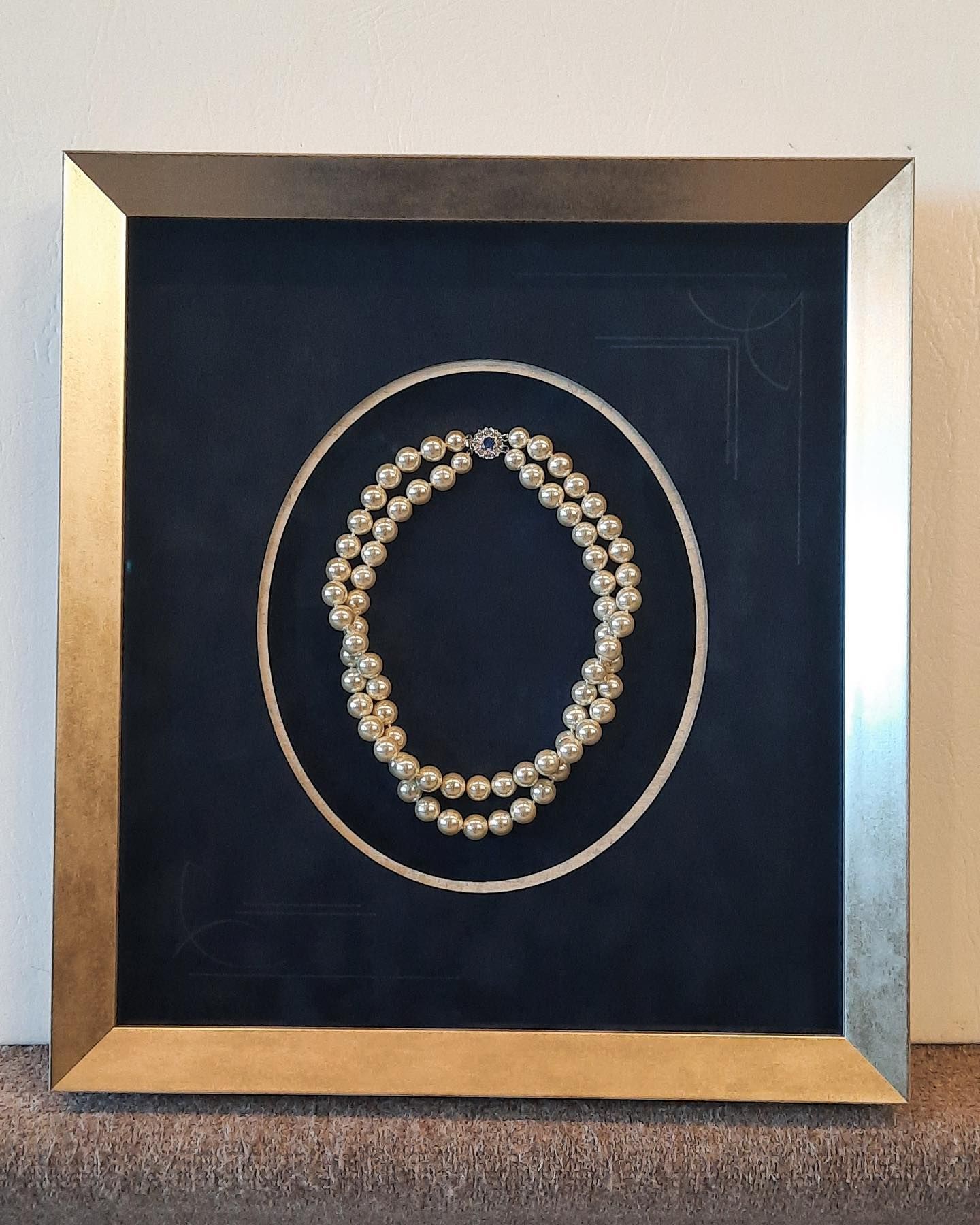 Framed pearl necklace on dark blue background, inside a gold-toned frame.