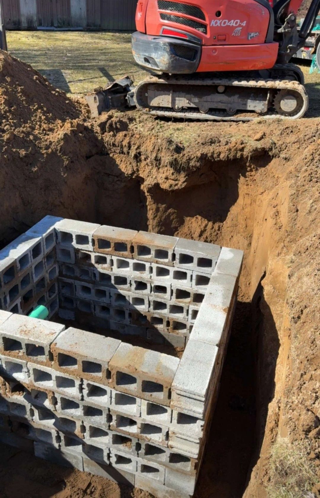 An excavator is digging a hole in the ground next to a brick wall.