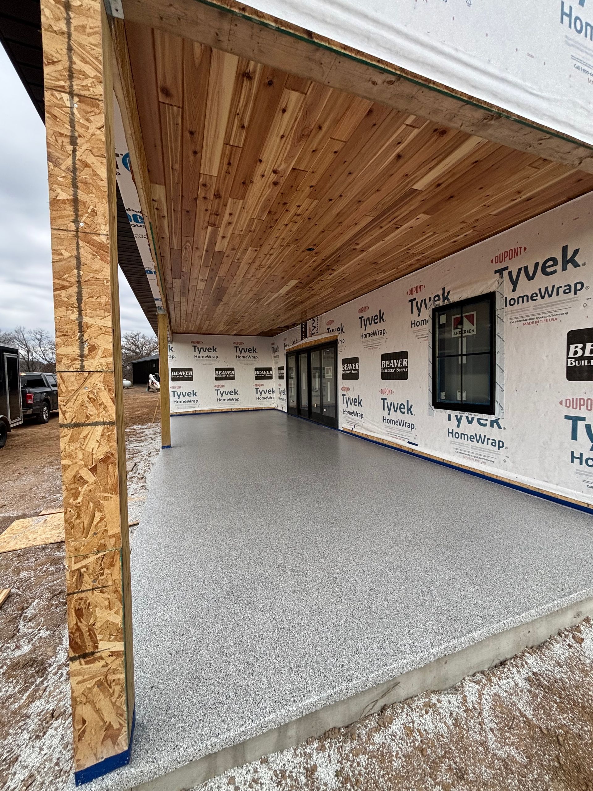 Partially built home entry with plywood ceiling and Tyvek-wrapped walls, opening to a gray concrete patio