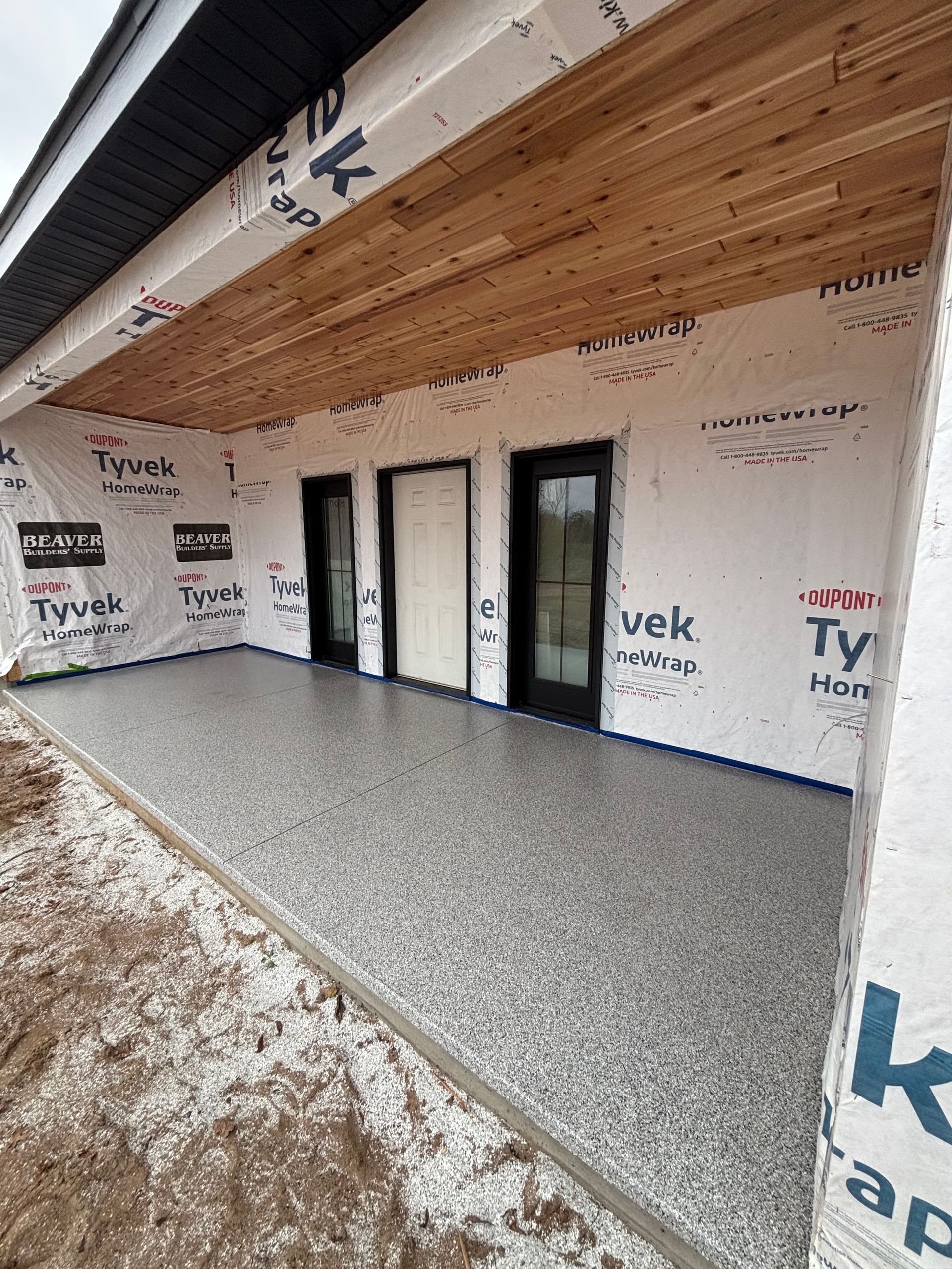 Covered porch under construction with unfinished wood ceiling, black-framed doors, and gravel ground.