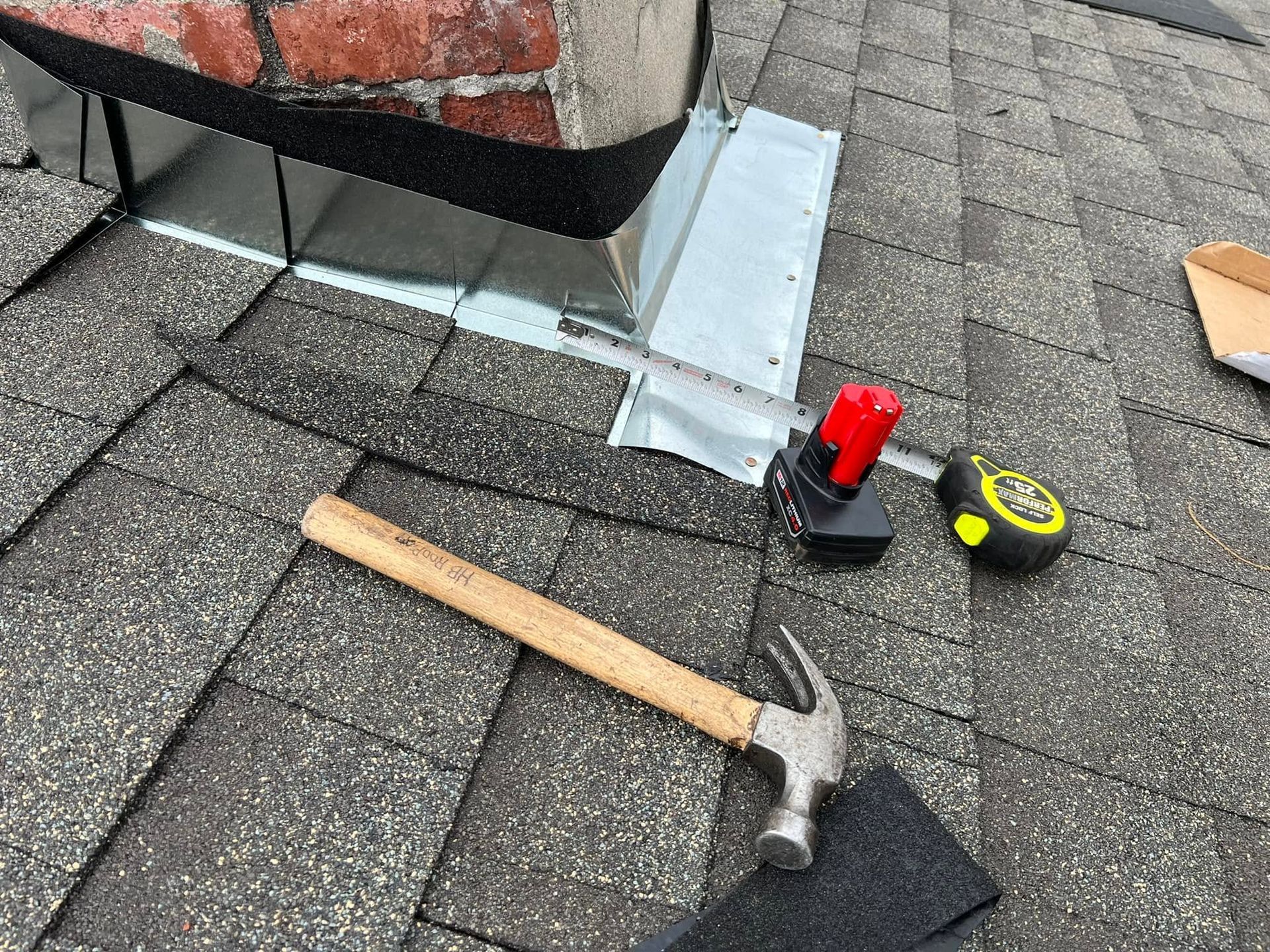 Roofing tools and a hammer on gray shingles beside a vent and metal flashing.
