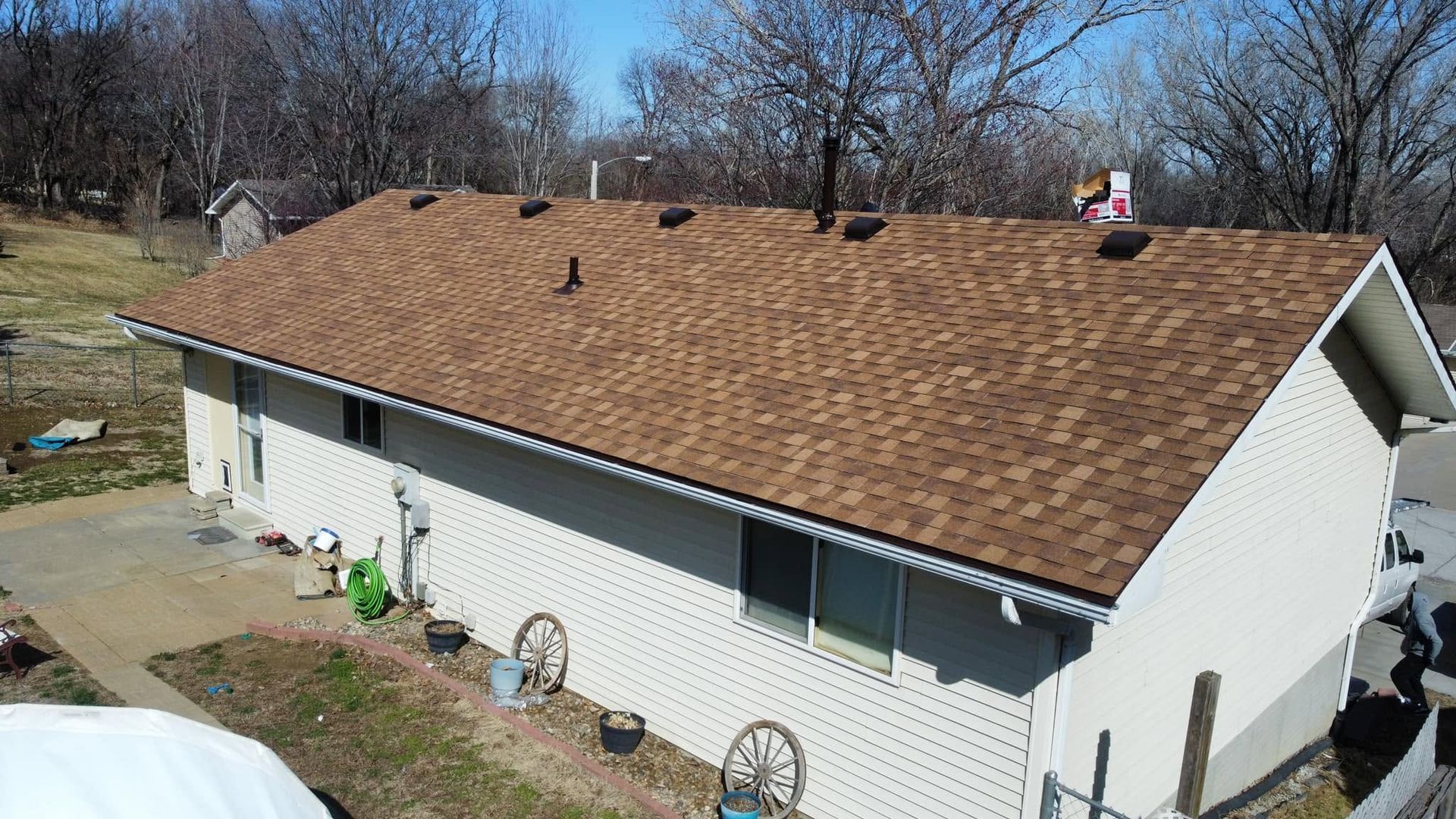 White house with a brown shingled roof, viewed from above in a yard with leafless trees.