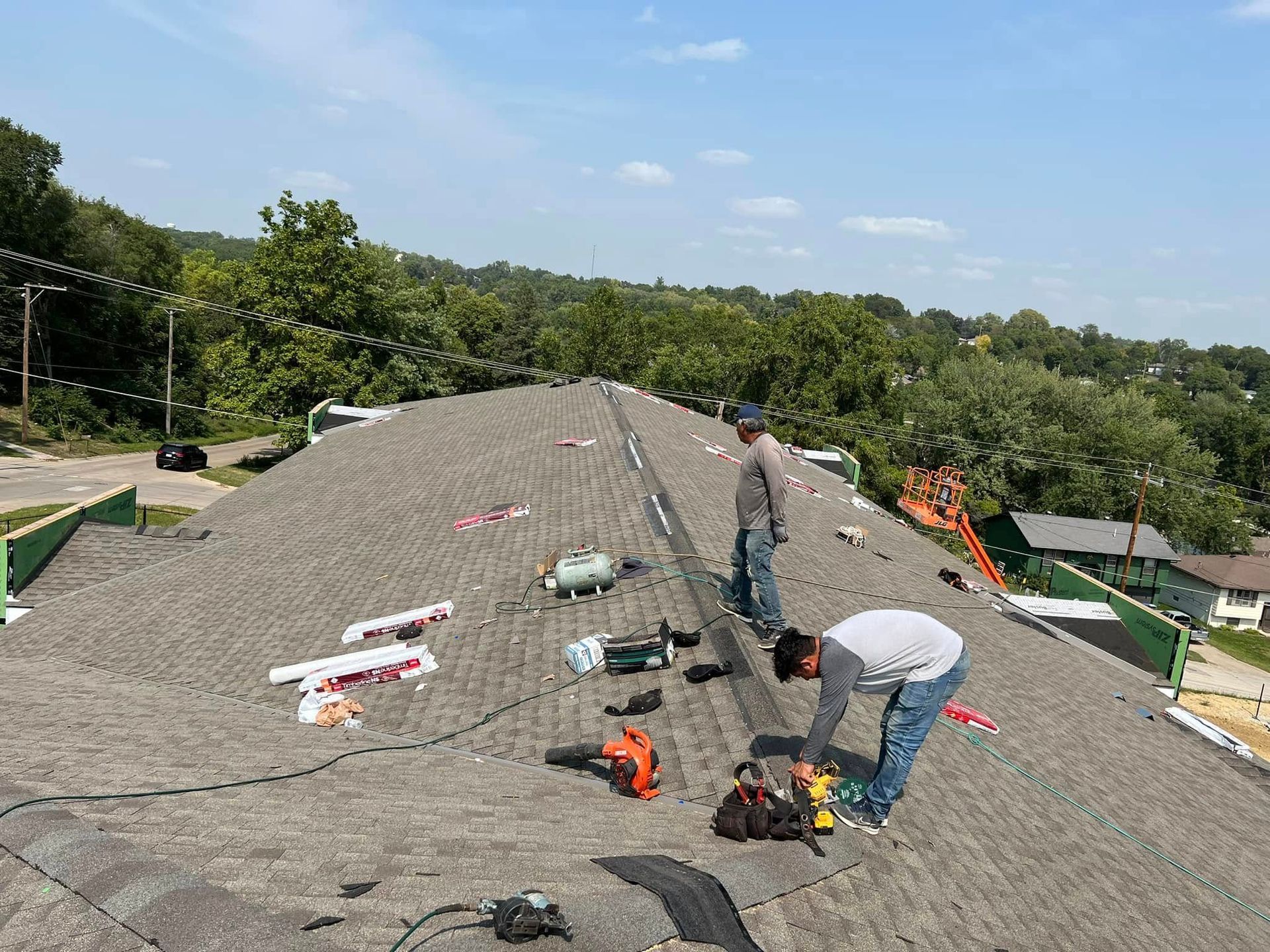 Workers installing roofing on a steep gray roof, with tools and materials spread out under a blue sky.