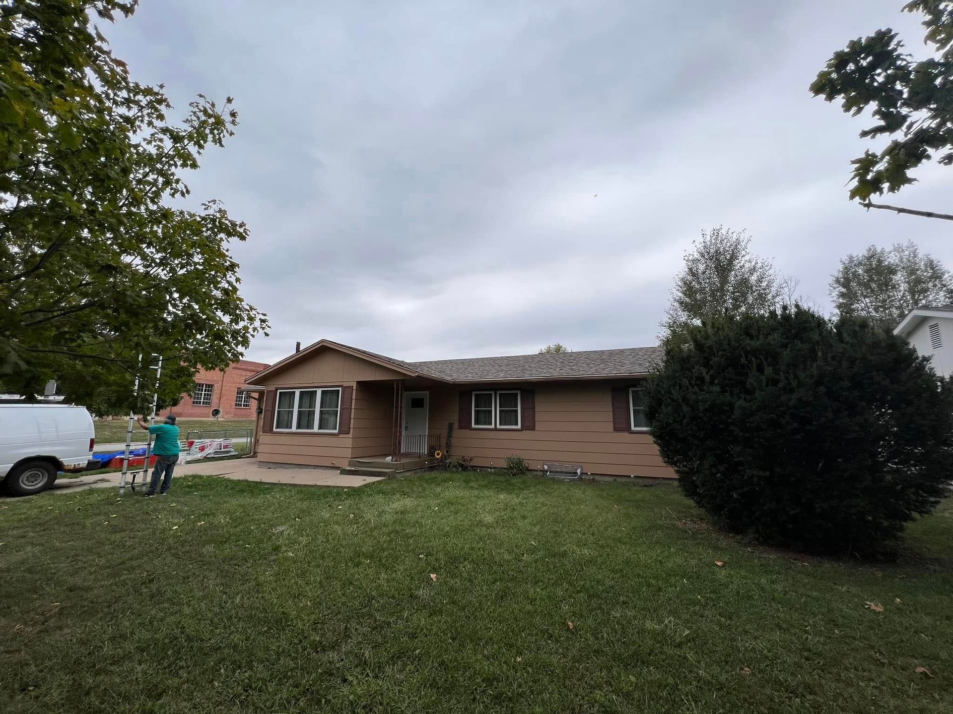 Single-story brick house with a front lawn, cloudy sky, and a person near a white van on the left.