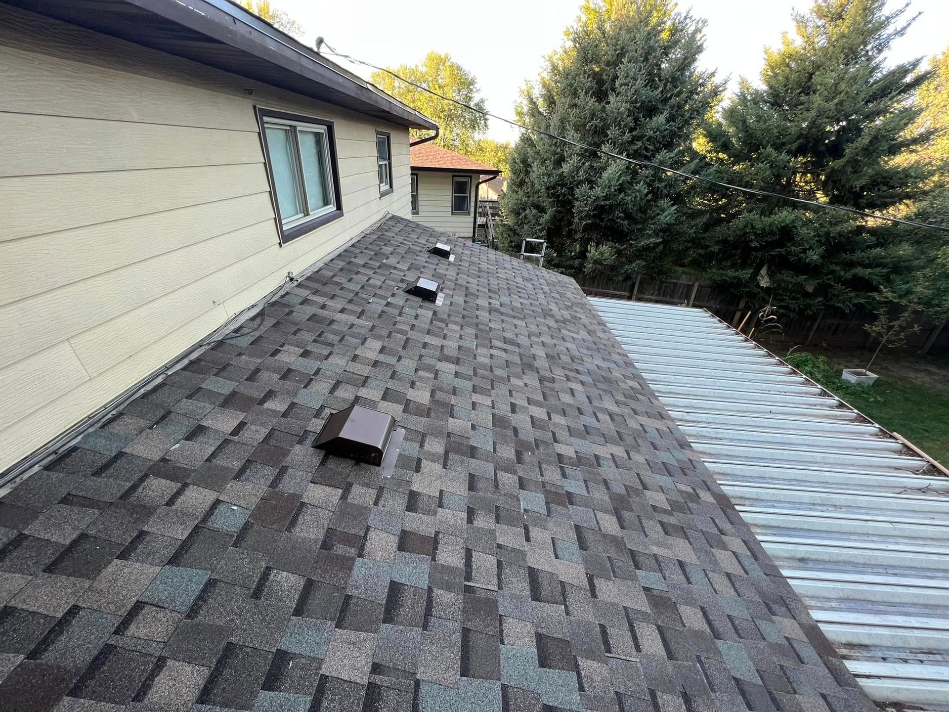 Roof with new gray shingles beside an older corrugated metal roof, viewed along a house exterior.