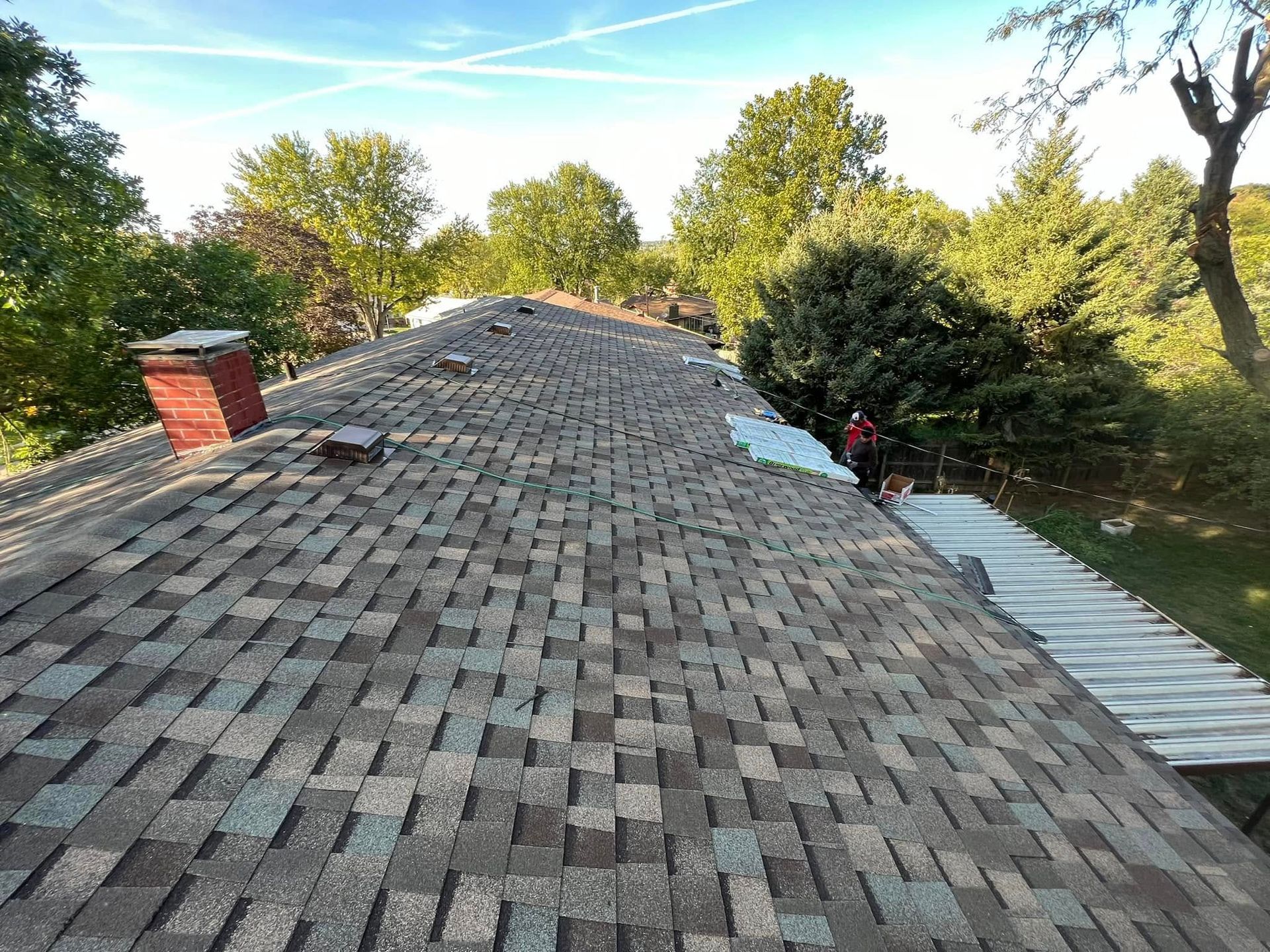 Roof shingles on a sloped house roof with trees and blue sky in the background