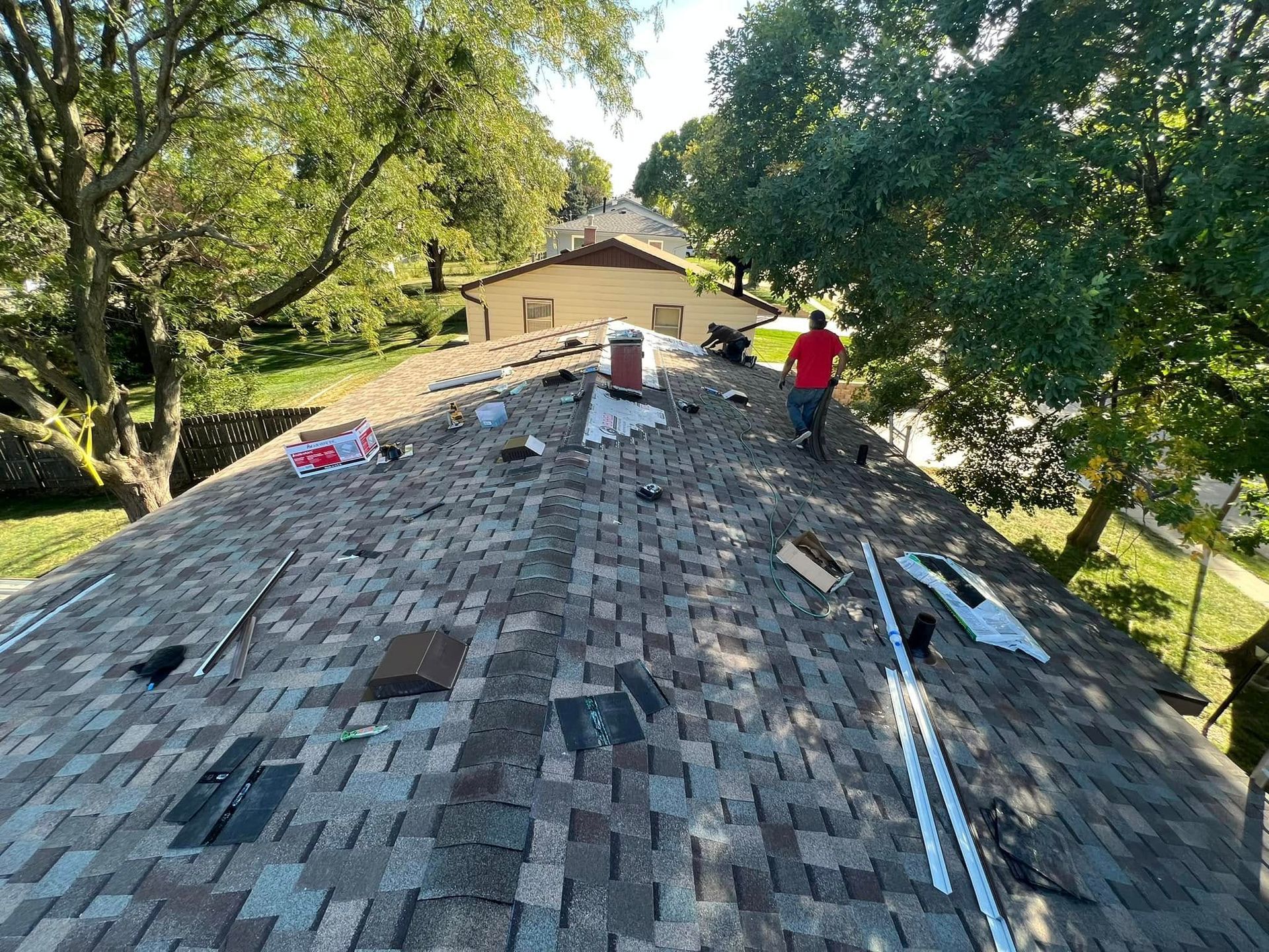 Roof under repair with two workers and scattered tools in a tree-lined yard