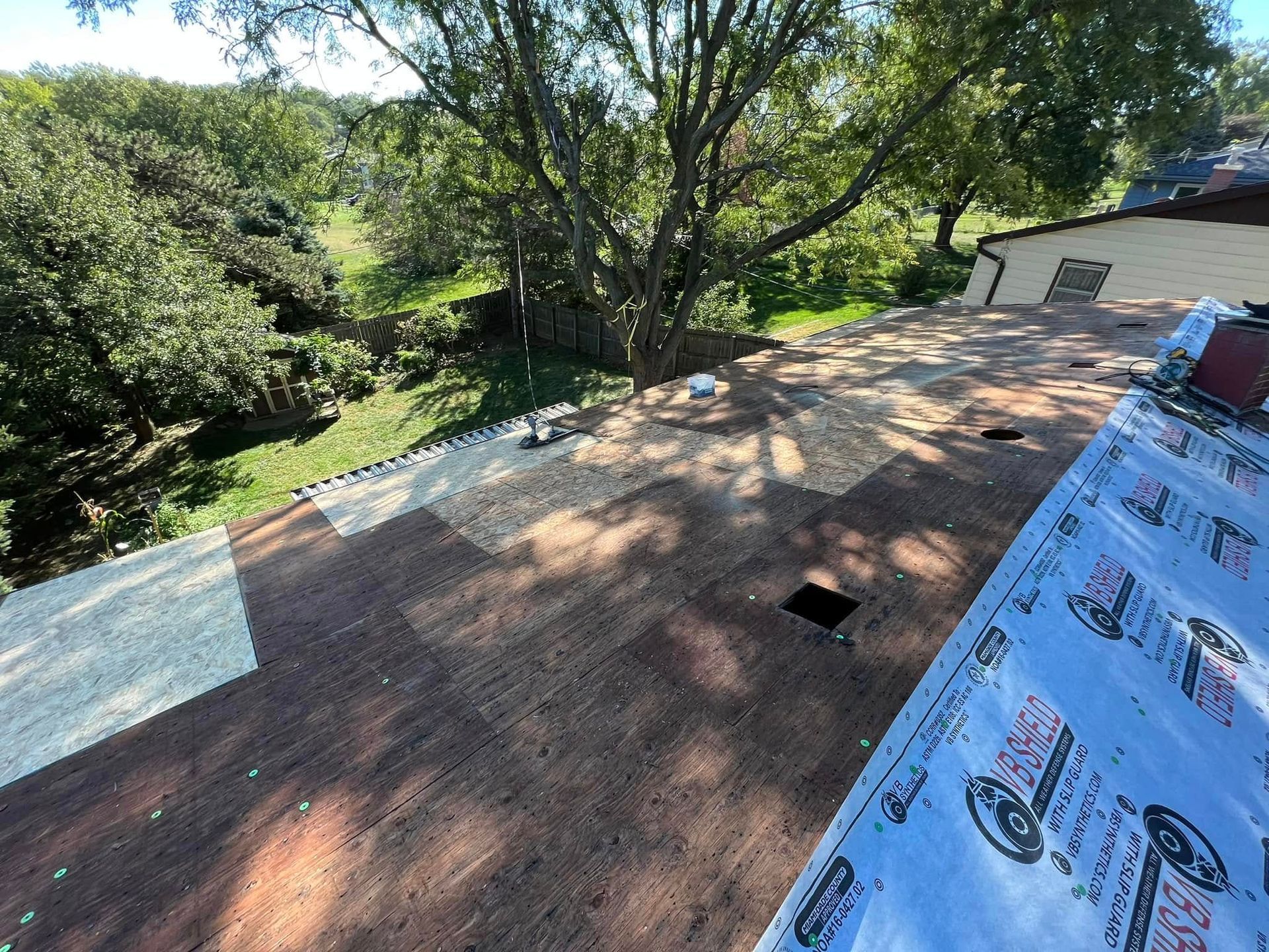 Roof under repair with exposed shingles and roofing felt, surrounded by trees in bright daylight