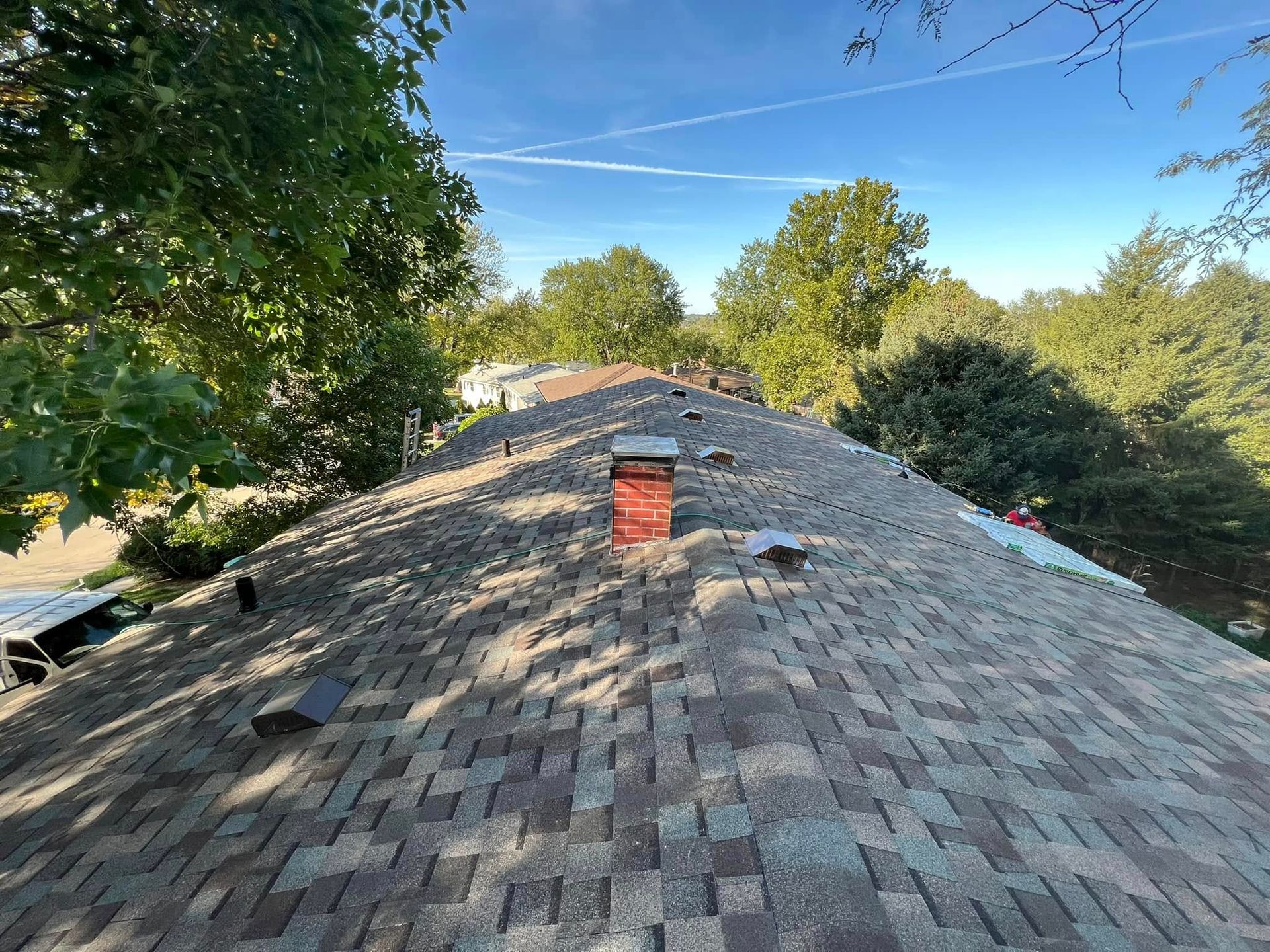 Shingled rooftop with a red chimney, surrounded by green trees under a clear blue sky.