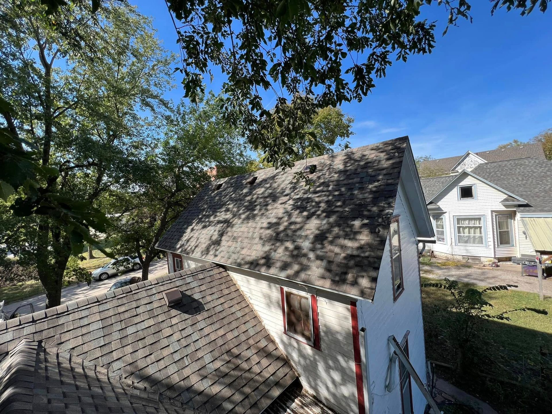 Small white shed with gray shingle roof under trees, beside a house on a sunny day