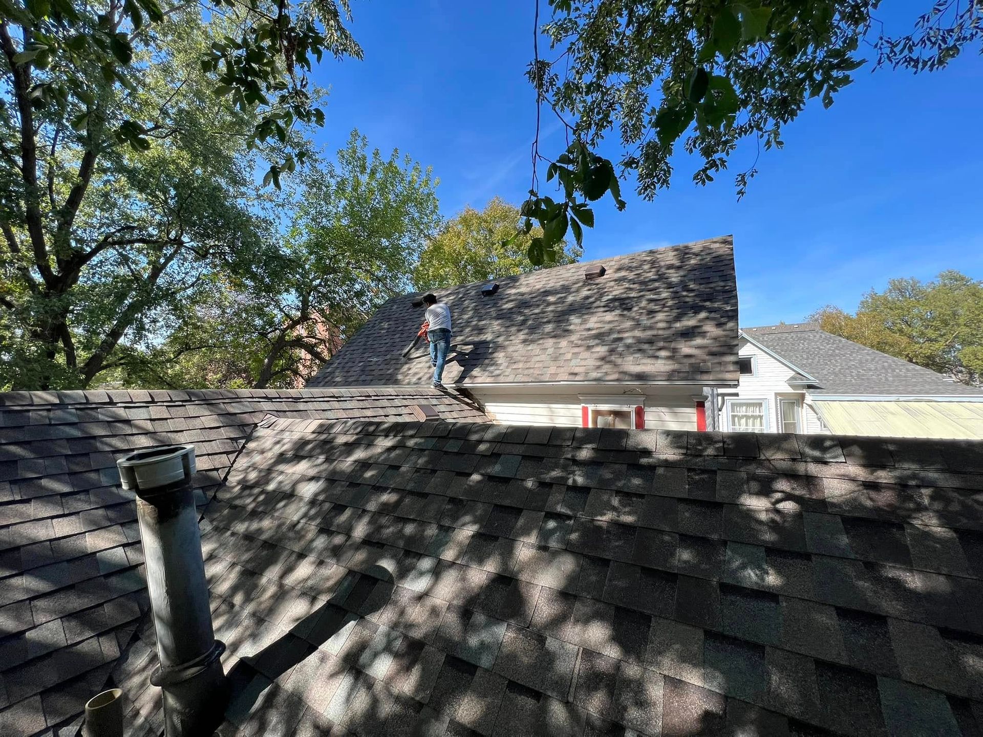 Roofer working on a shingled house roof under a blue sky with trees nearby
