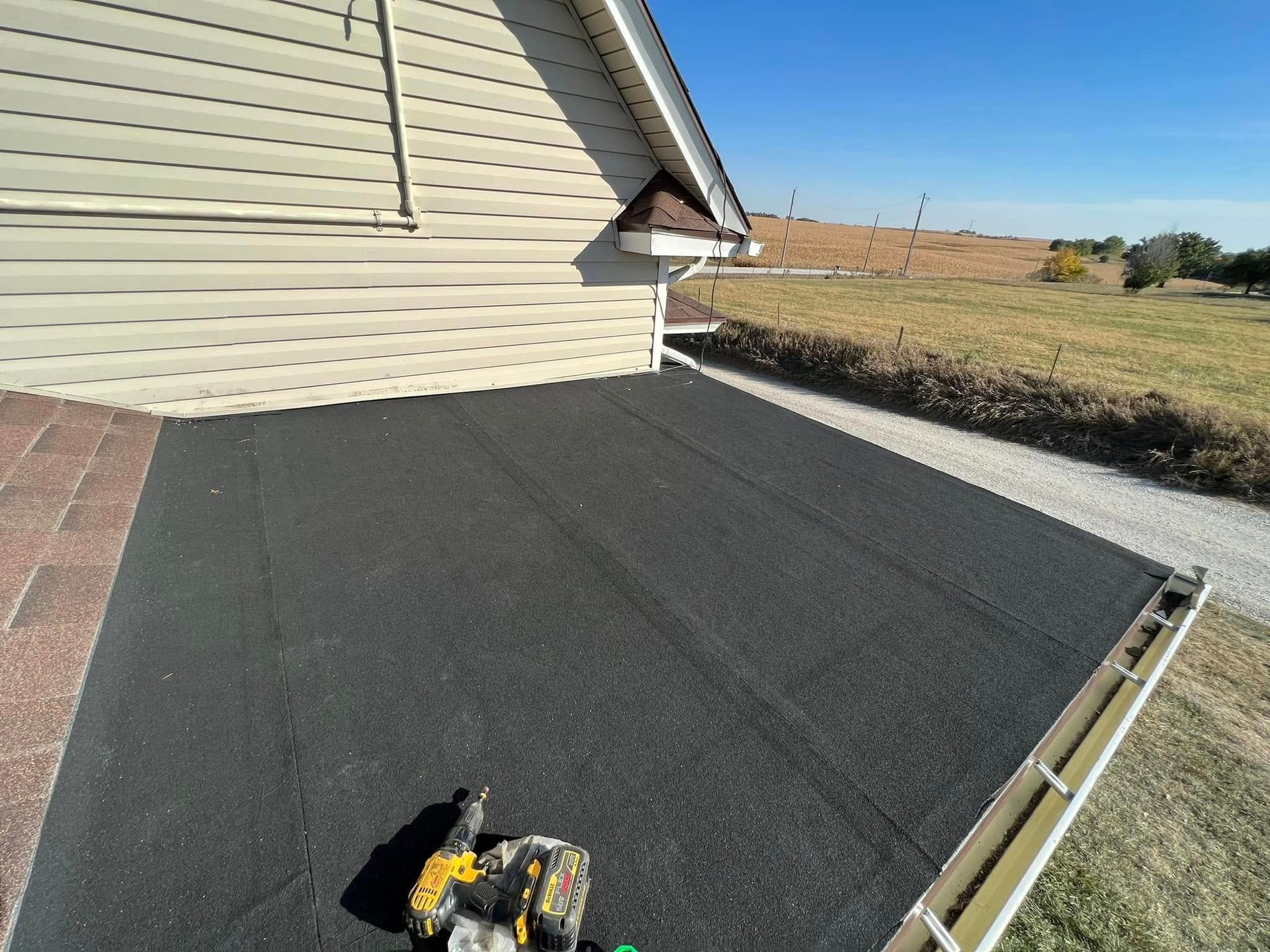 Black roofing membrane installed beside beige house wall, with tools on the roof in a rural yard.