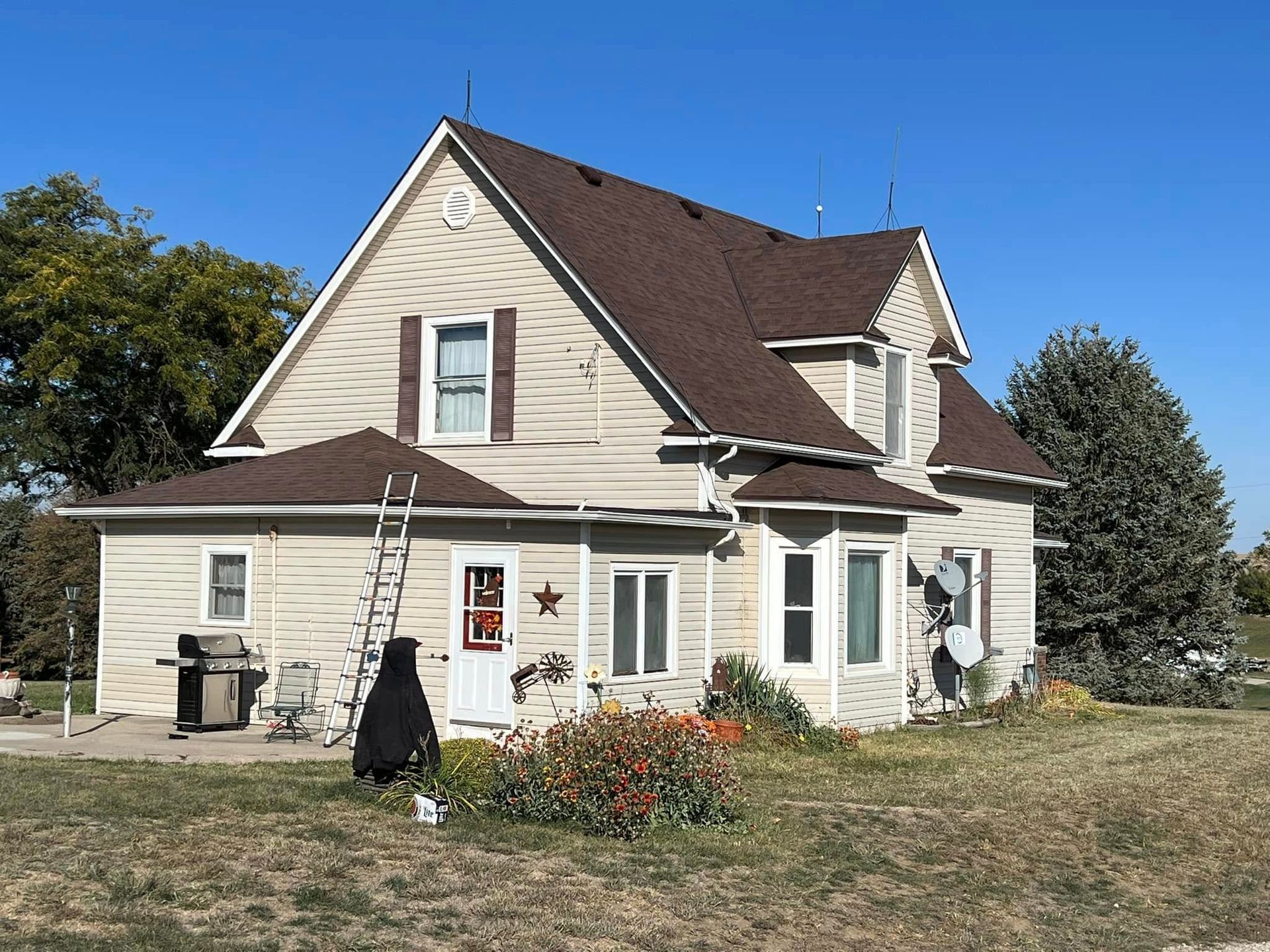Small beige house with a brown roof, ladder at the window, and flowers in the front yard.