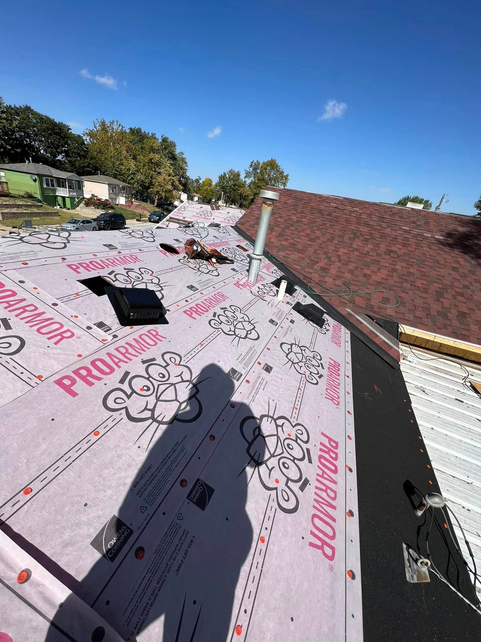 Roof under repair with pink underlayment, shingles, tools, and a worker’s shadow under a blue sky