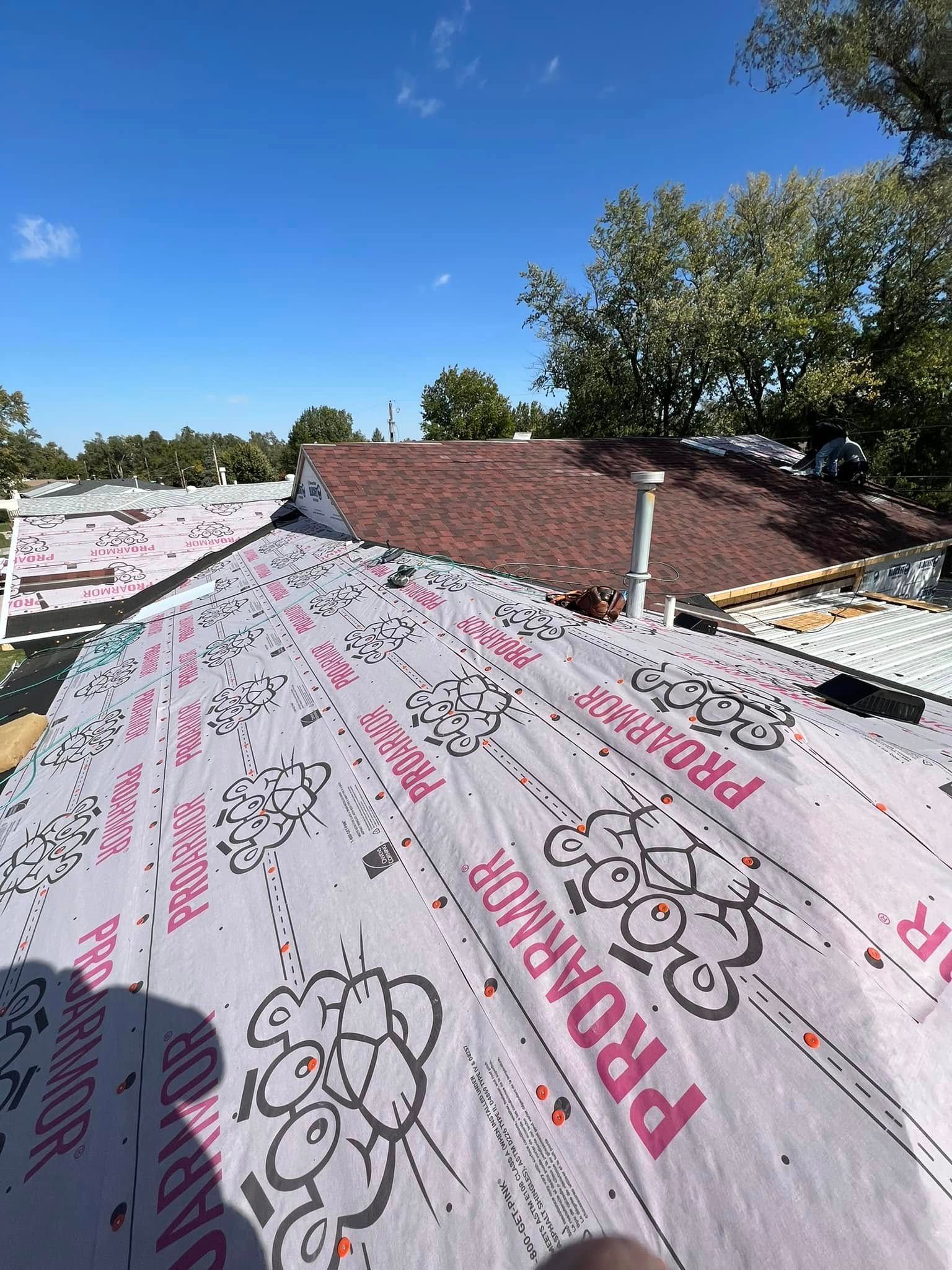 Rooftop underlayment with pink printed membrane beside a brown shingled roof and trees under a blue sky