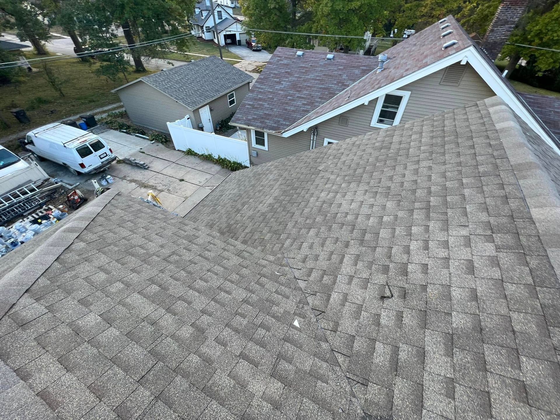 View from a rooftop over gray shingles to neighboring houses, trees, and a parked white van