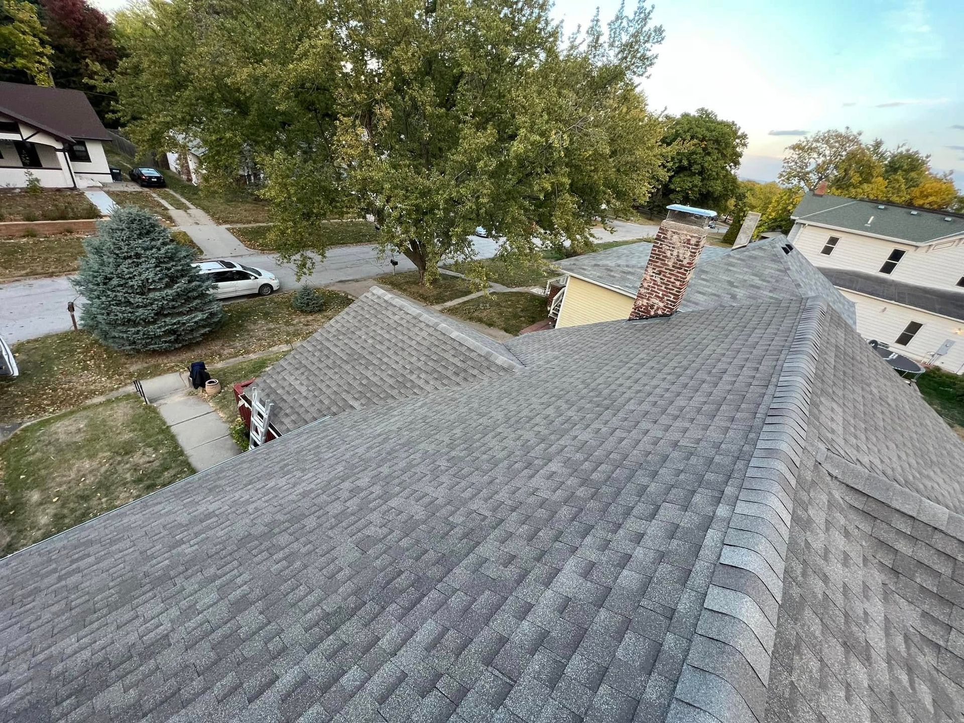 View from a gray shingled roof over a tree-lined suburban neighborhood with houses and a chimney.
