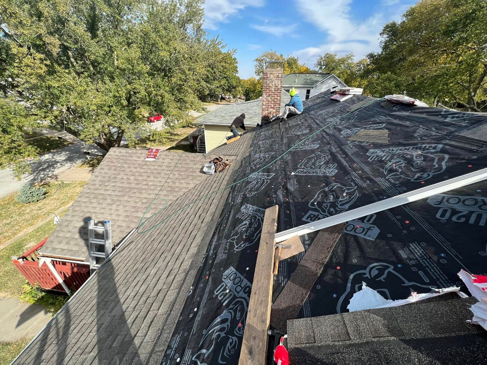 Roofers working on a shingled roof with black underlayment on a tree-lined house roof.